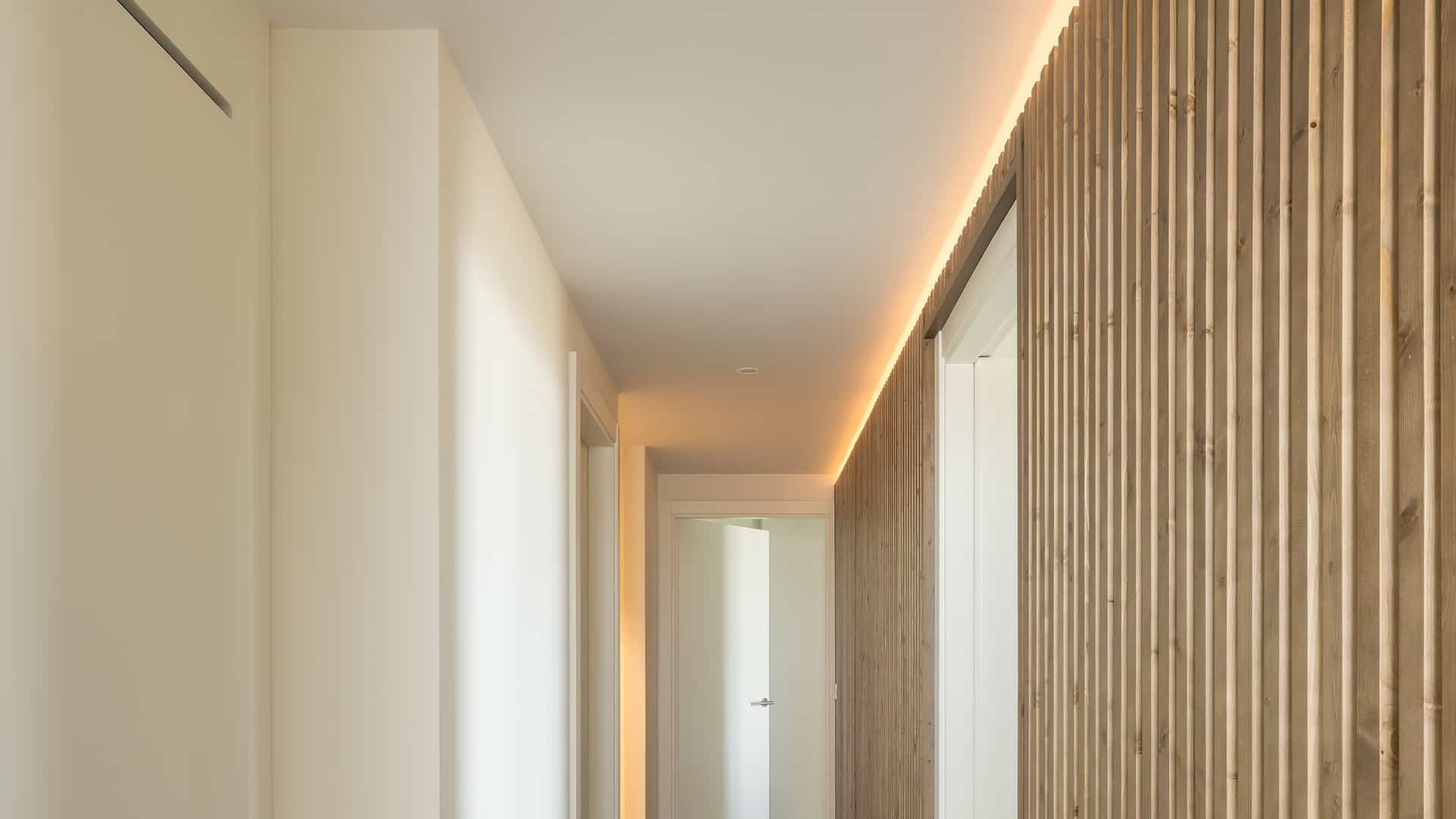 Pasillo con cornisa de luz lineal LED continua sobre pared de madera, proyecto residencial JISO